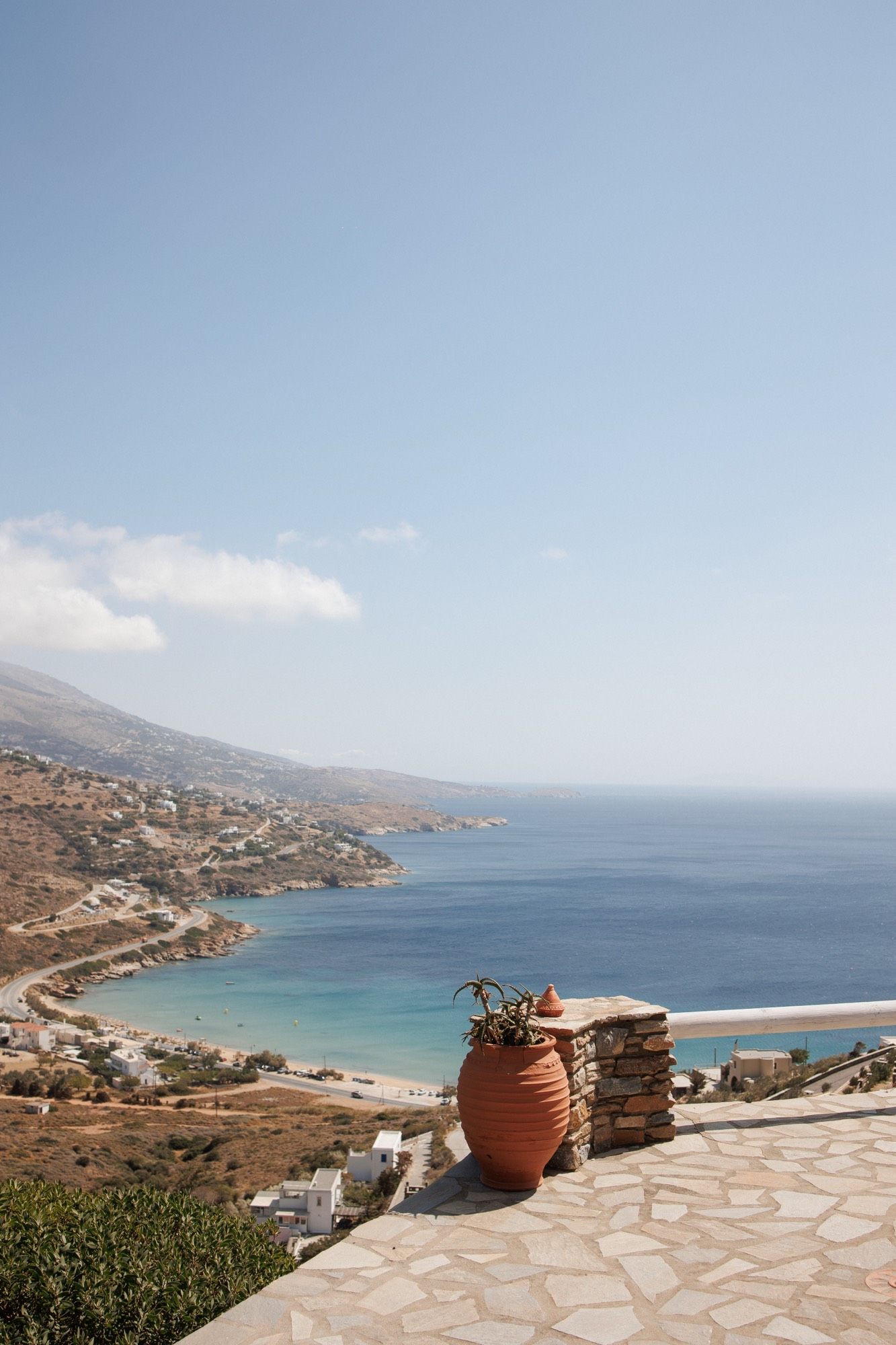 Panoramic coastal views of a Mediterranean bay with terracotta pots on stone patio overlooking turquoise waters and hills.