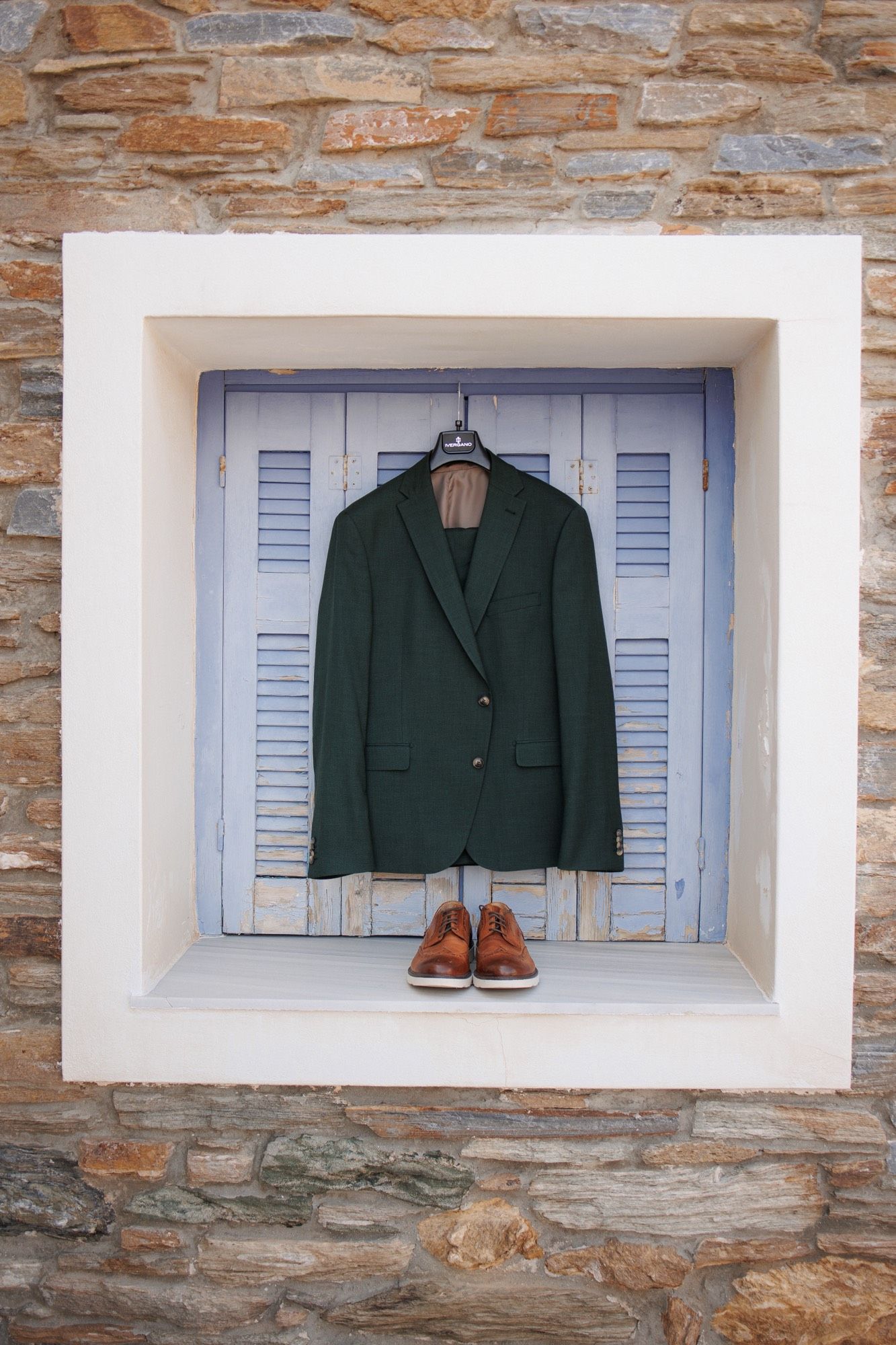 A green blazer and brown leather shoes displayed in white window alcoves against a rustic stone wall.