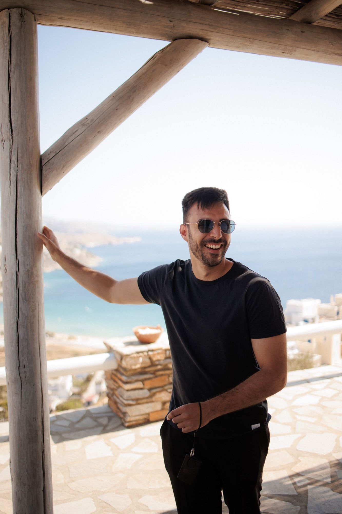 Person wearing sunglasses and black shirt smiles while leaning on white beams with ocean view in background.