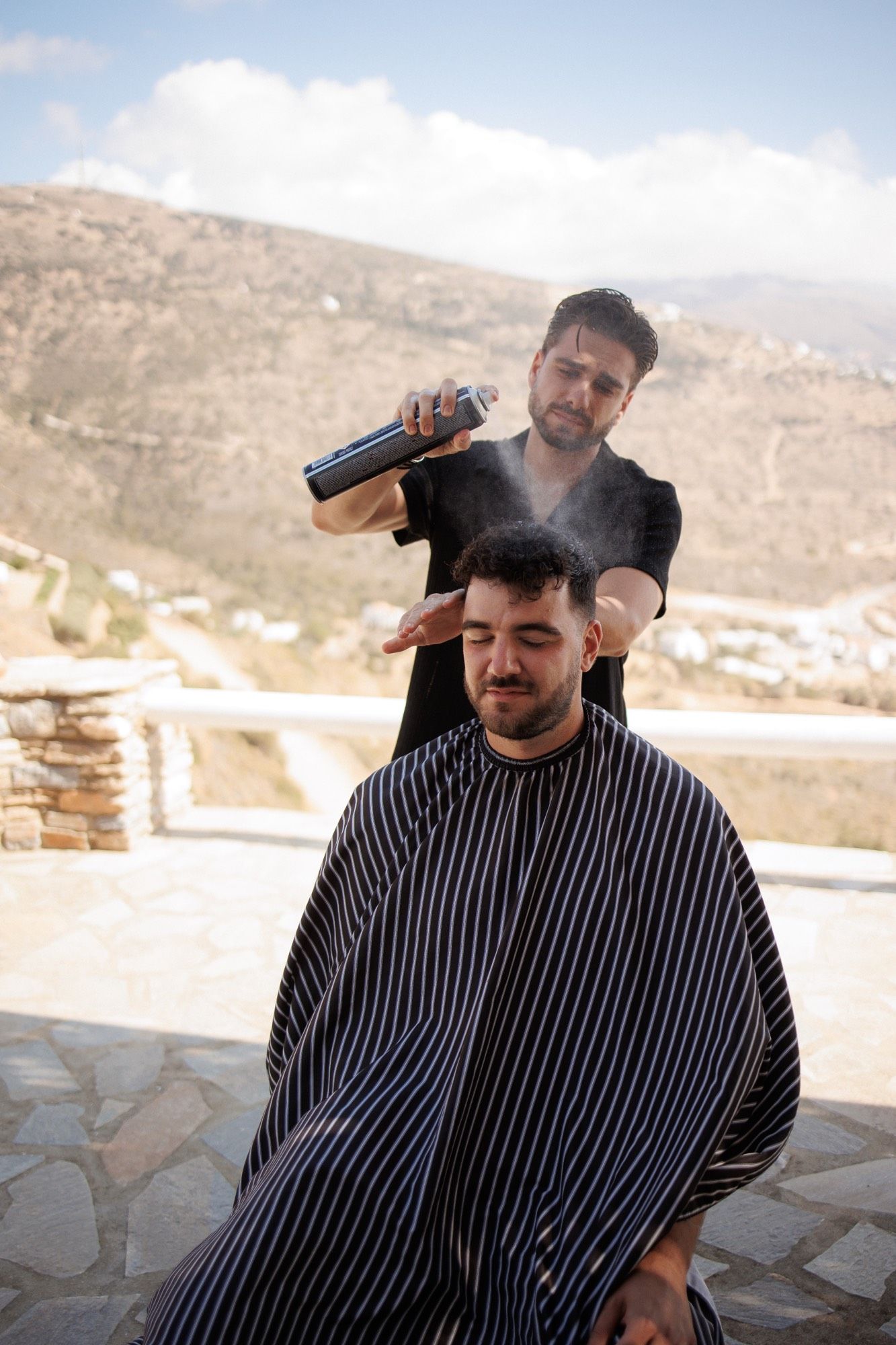 A barber in a striped cape provides an outdoor haircut service with mountains visible in the background.