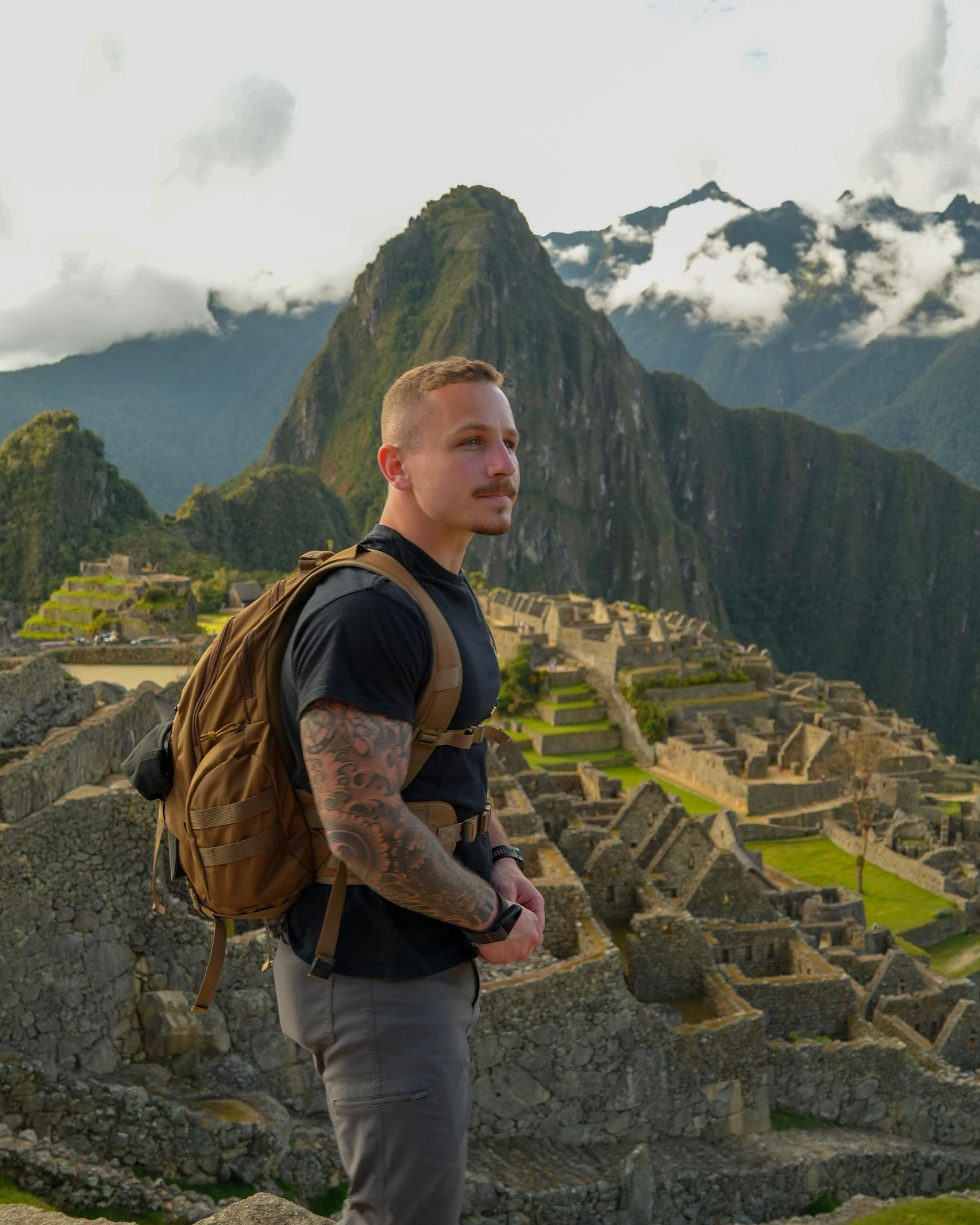 Tattooed man with backpack standing at Machu Picchu ruins with mountains in the background.