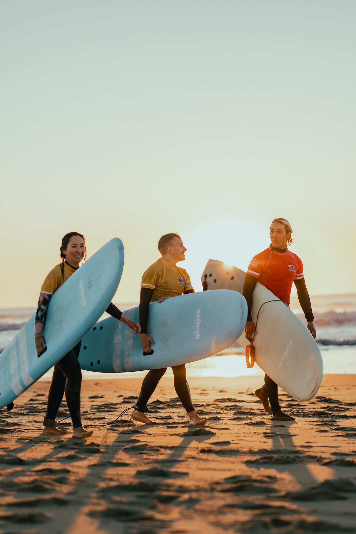 Des surfeurs portant des planches de surf marchant le long d'une plage de sable au coucher du soleil dans une séquence de plans d'action en