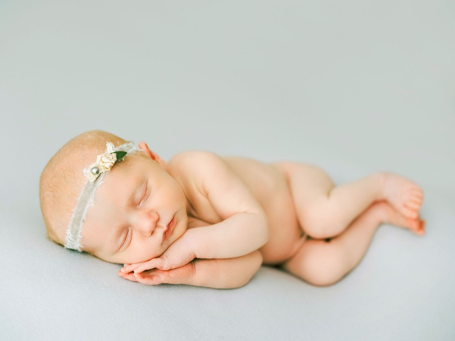 Peaceful sleeping newborn baby wearing delicate white headband lying on soft gray backdrop.