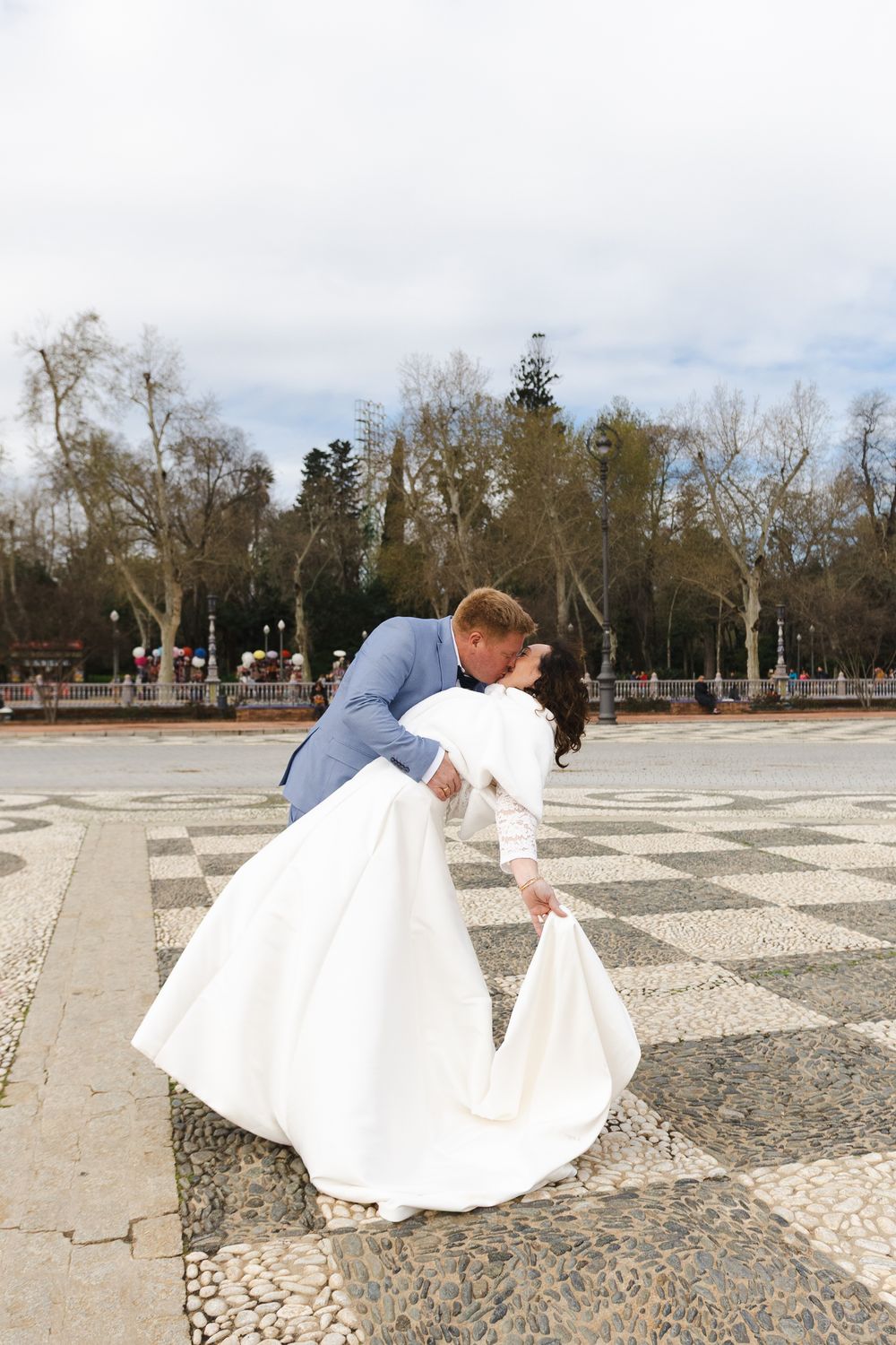 A couple in wedding attire dances gracefully on a stone patterned courtyard with trees in the background.