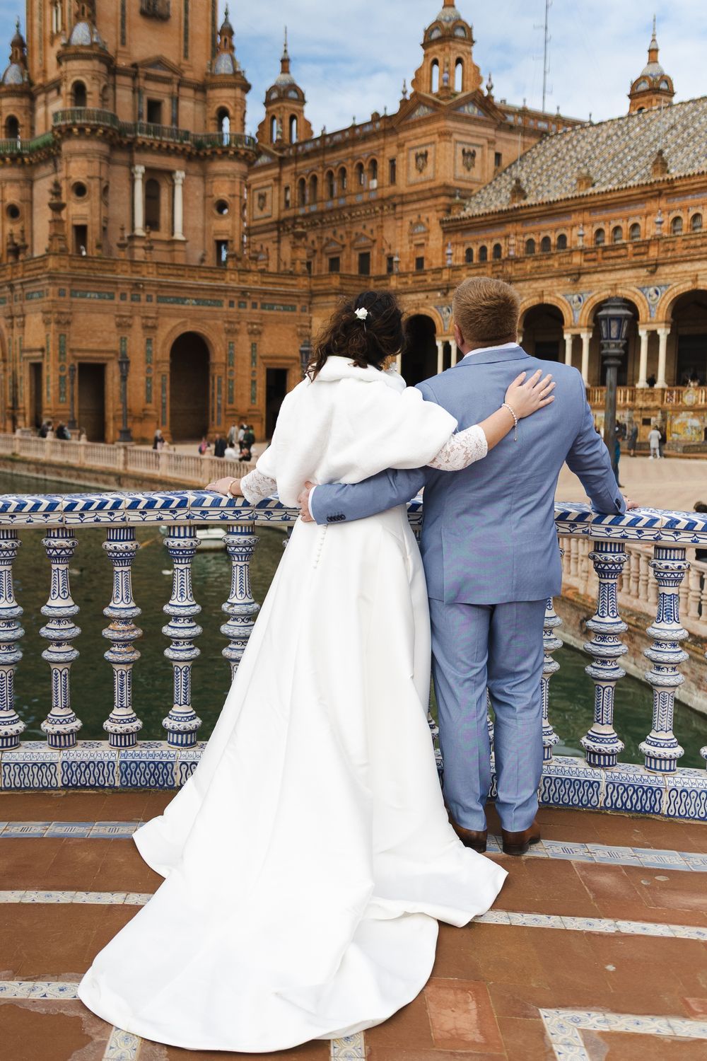 A couple in wedding attire stands at the ornate Plaza de Espana in Seville, Spain, with its grand architecture in the background.