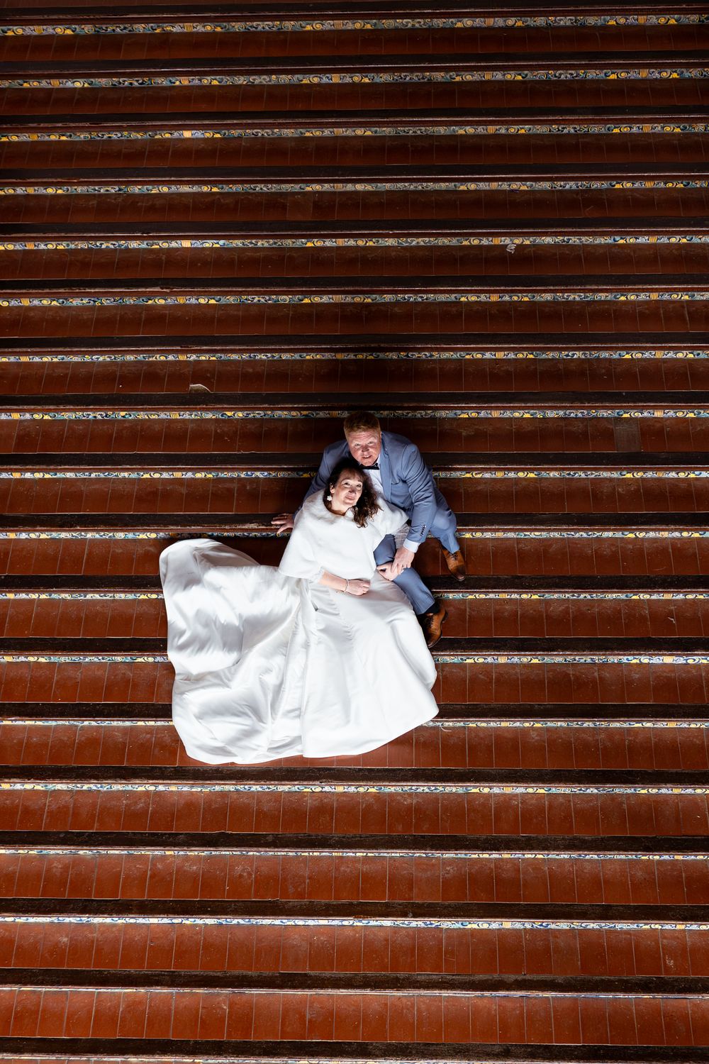 A couple in wedding attire lies artistically across red brick stairs in a creative photo composition.