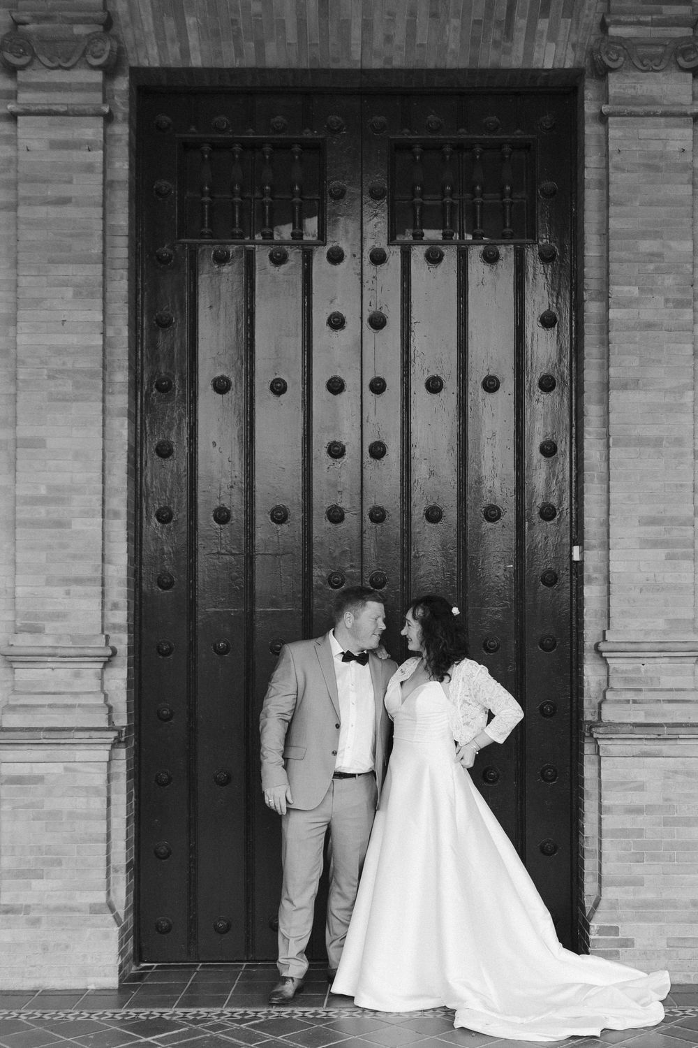 A romantic couple in wedding attire poses in front of a grand wooden door with ornate metalwork details.