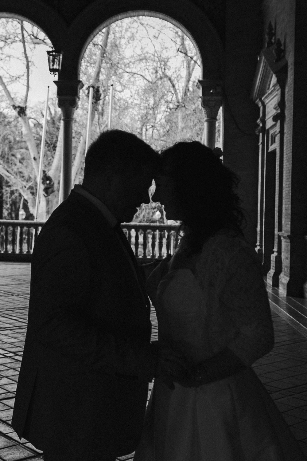 Three black and white silhouettes of people stand under a covered porch with trees visible in the background.