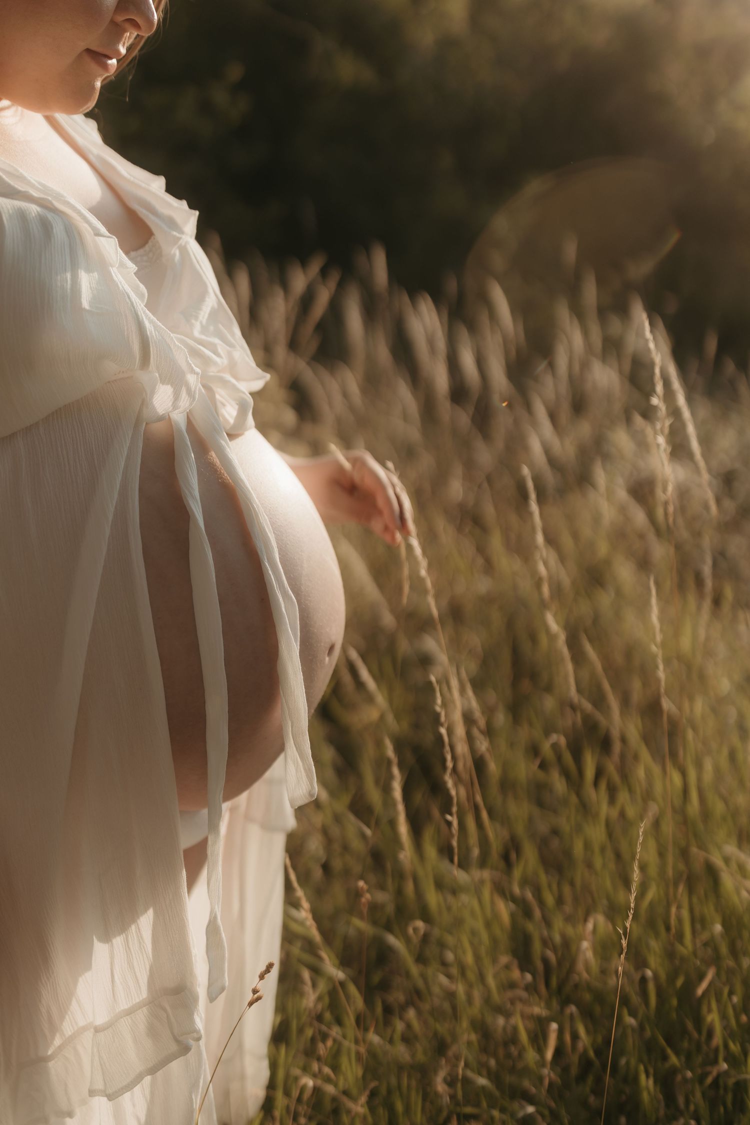 A person in a flowing white dress stands in a sunlit meadow of tall grass at golden hour.