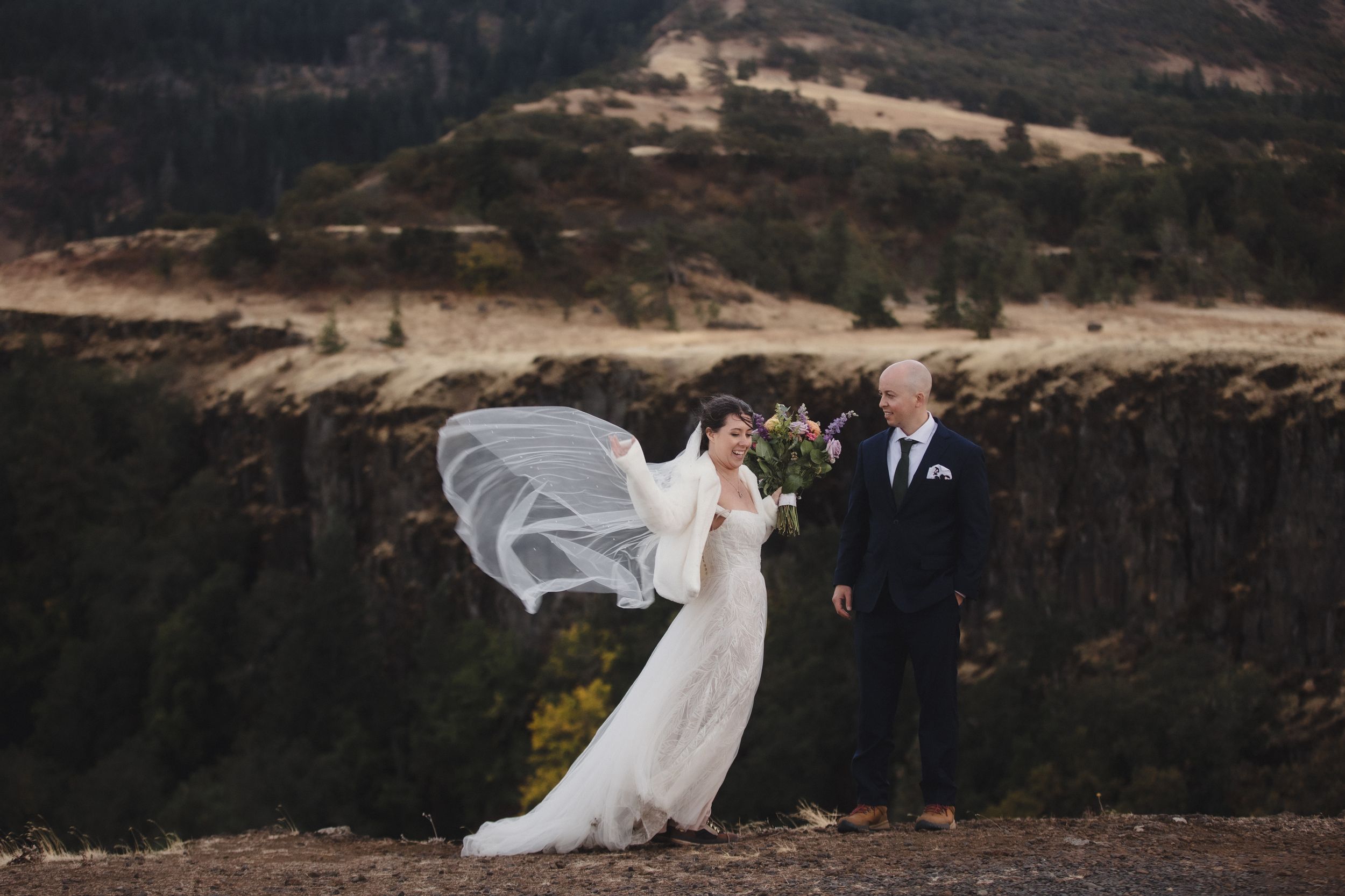 A wedding couple poses on a mountain ridge as a flowing white veil catches the wind in dramatic fashion.
