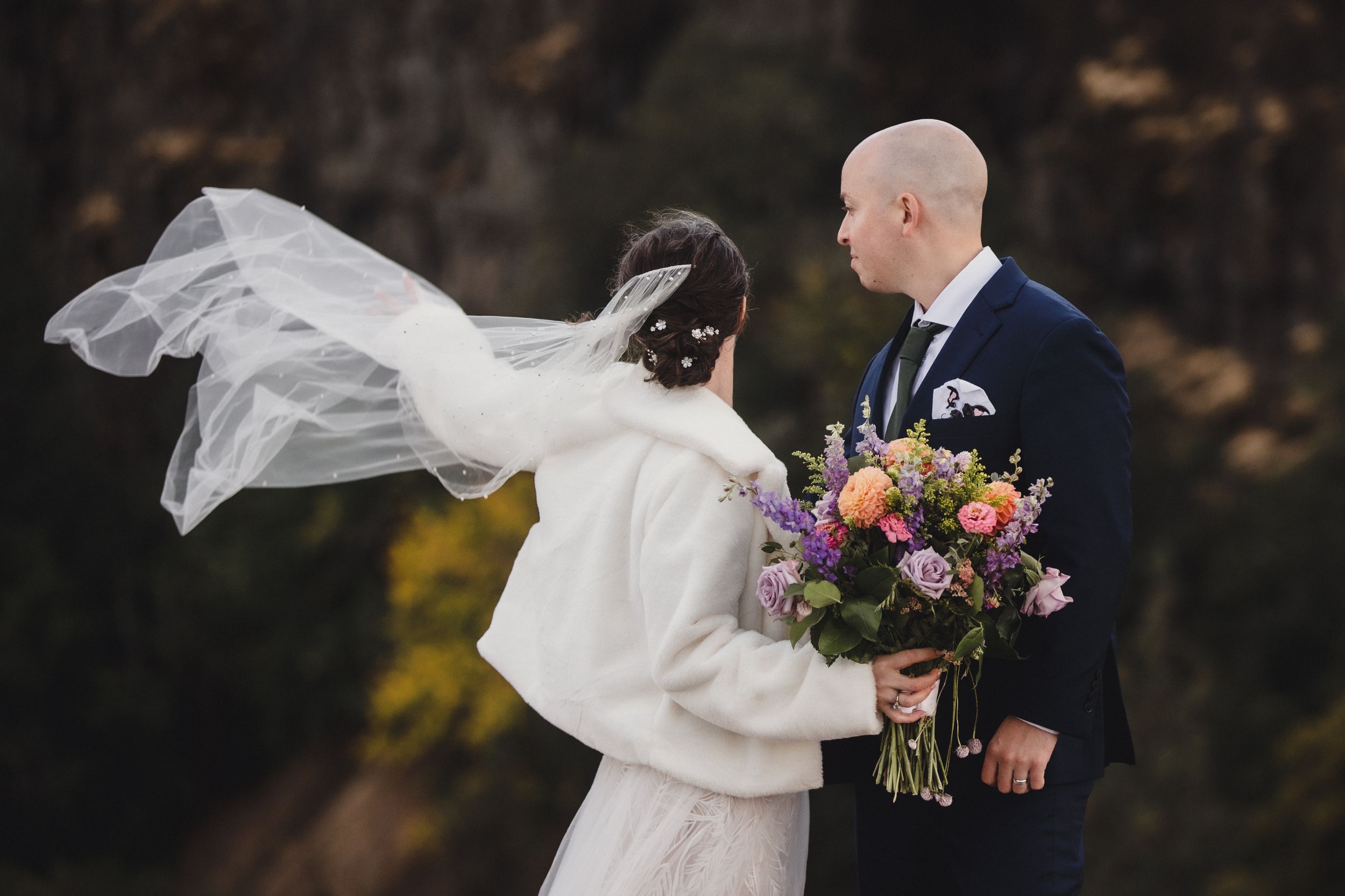 A bride's veil flows dramatically in the wind as she holds a colorful bouquet during an outdoor wedding photoshoot.