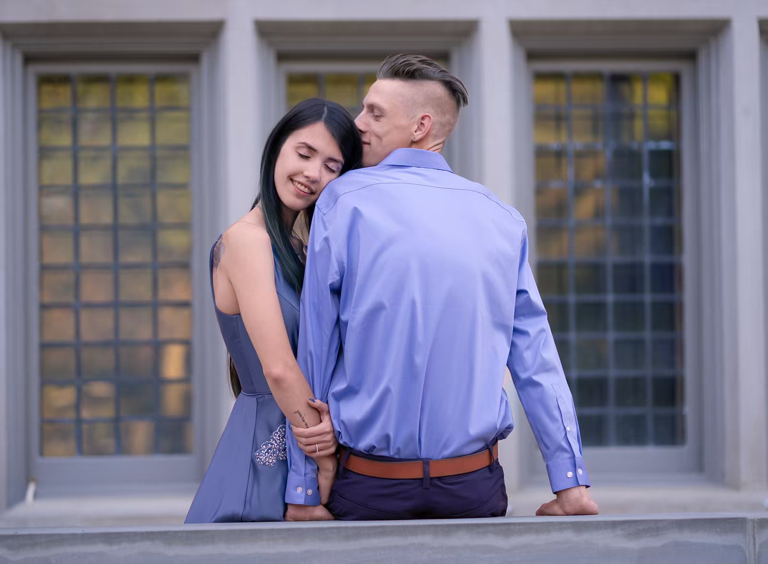 A couple poses against an architectural backdrop with large windows.