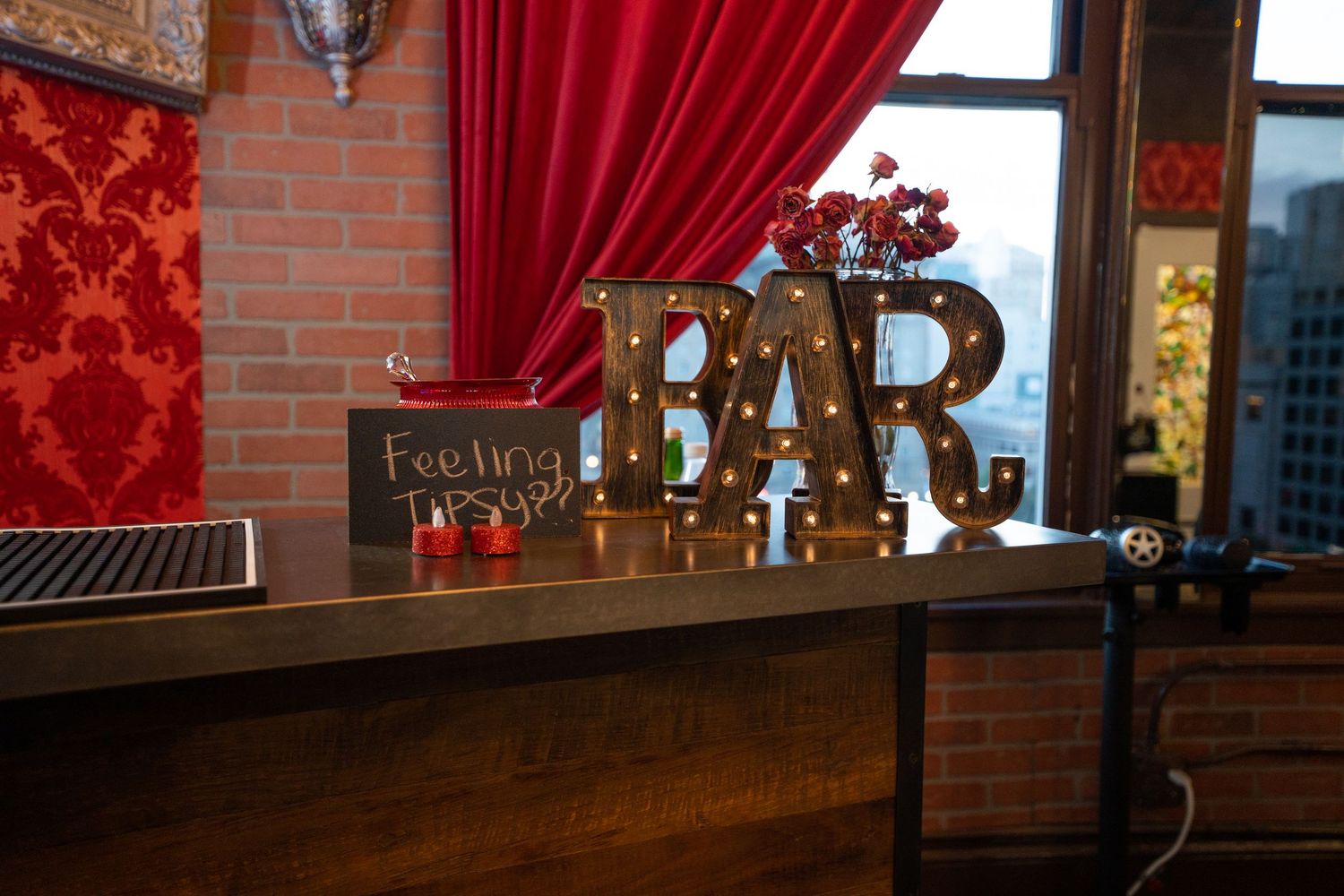 Illuminated gold BAR letters sit on a wooden counter against red damask wallpaper and exposed brick in a cozy lounge setting.