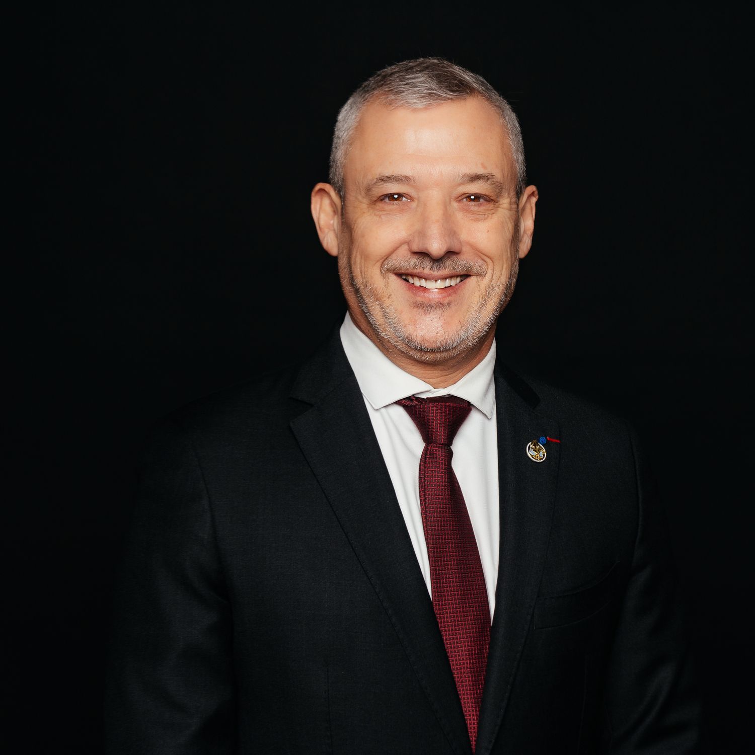 Professional man in black suit and burgundy tie smiling against dark background.
