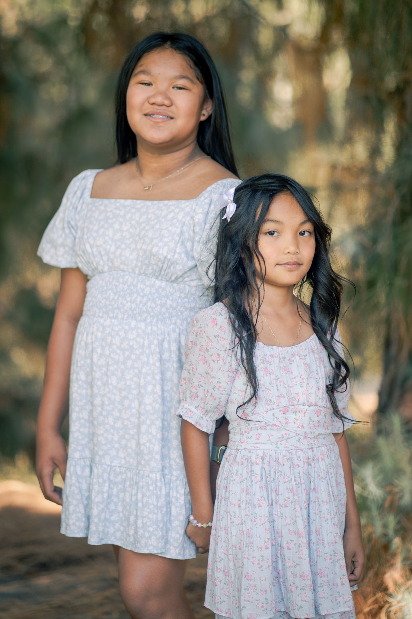 Two children wearing matching white floral dresses pose together in a wooded outdoor setting.