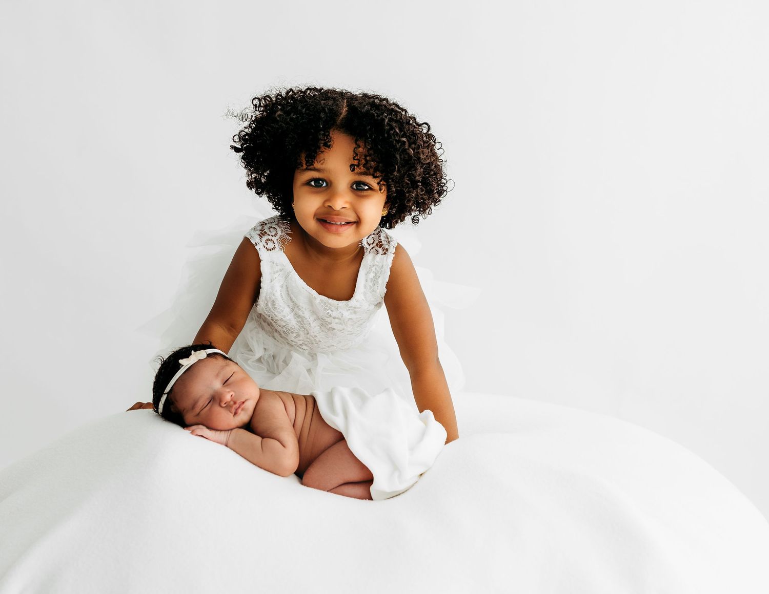 Toddler in white clothing gently interacting with sleeping newborn sibling on soft white bedding.