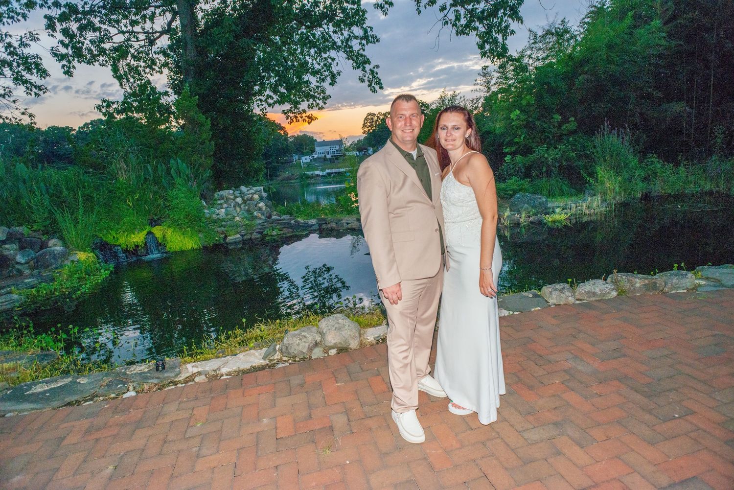 A couple in white outfits poses together in a romantic garden setting at sunset near a pond.