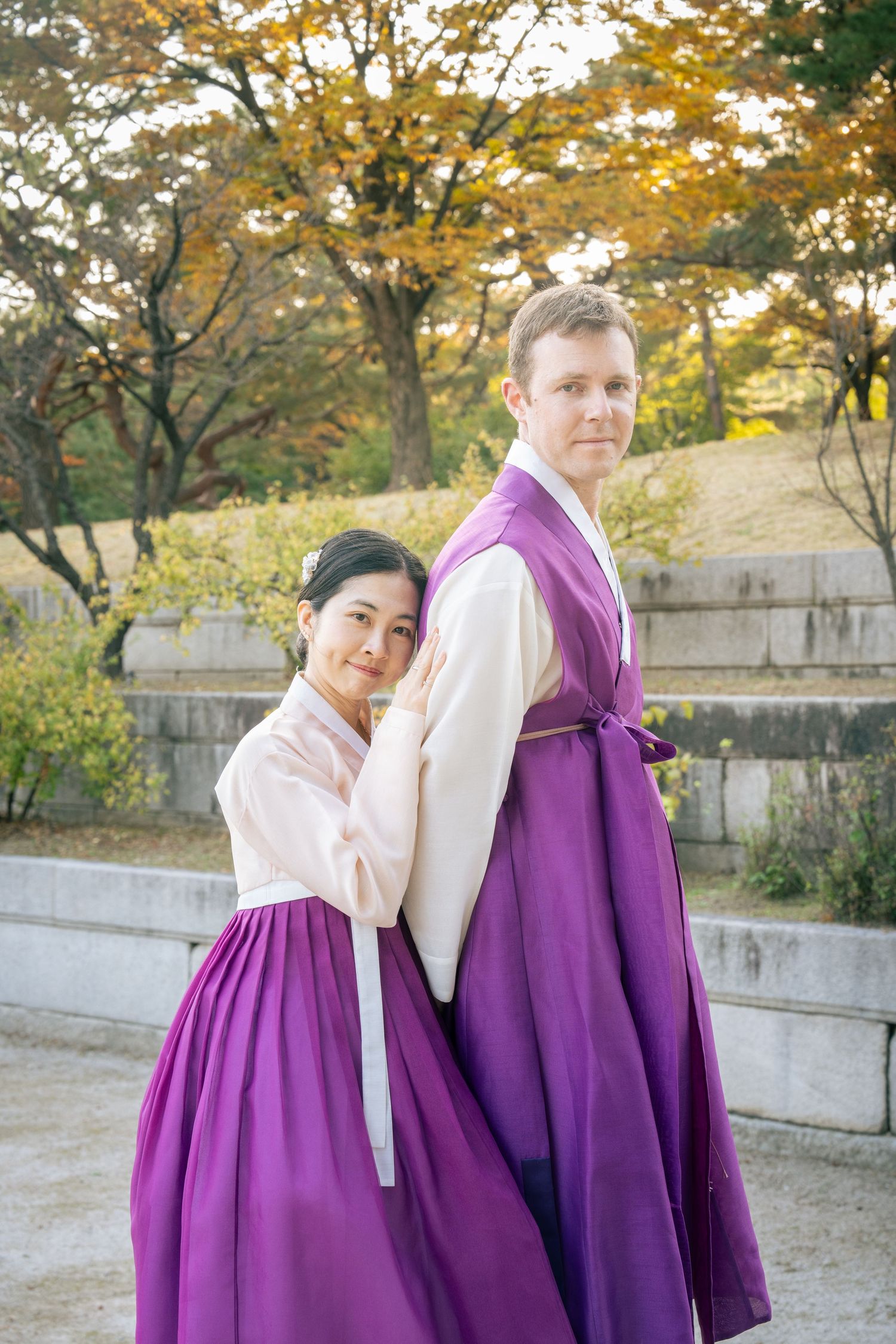 A couple in matching purple traditional Korean hanbok outfits poses together in an autumn garden setting.