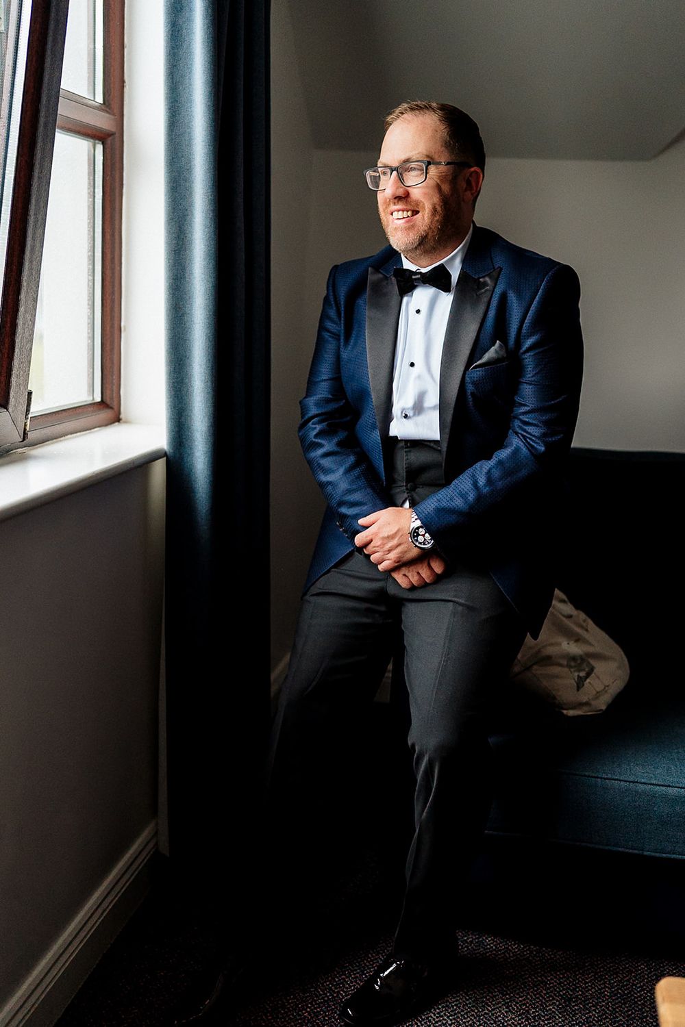 Person in navy blue tuxedo and bow tie sits casually by window in moody lighting.