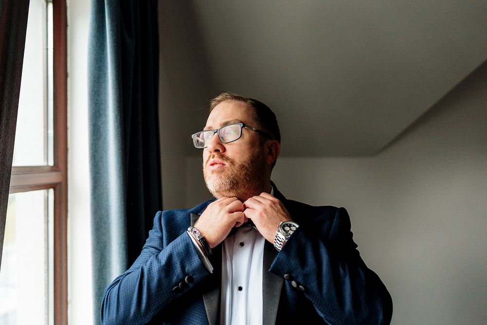 A formal-dressed person in glasses and navy suit stands near a window in natural light.