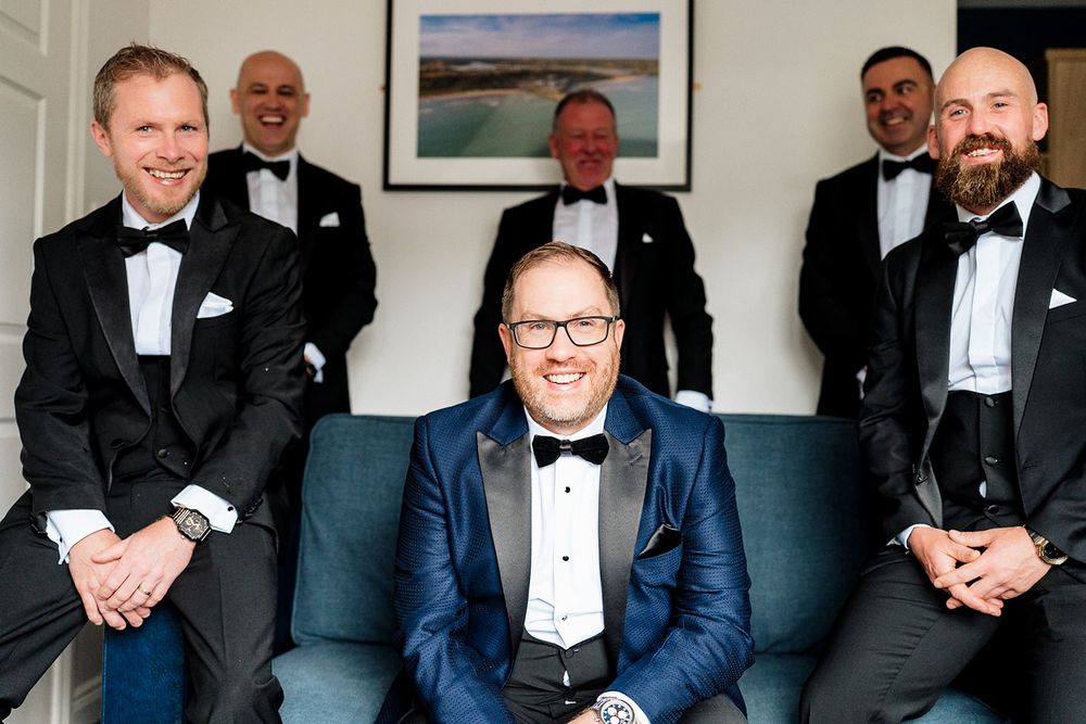 A formal event with attendees in black and navy tuxedos standing together in a room with framed artwork on the wall.