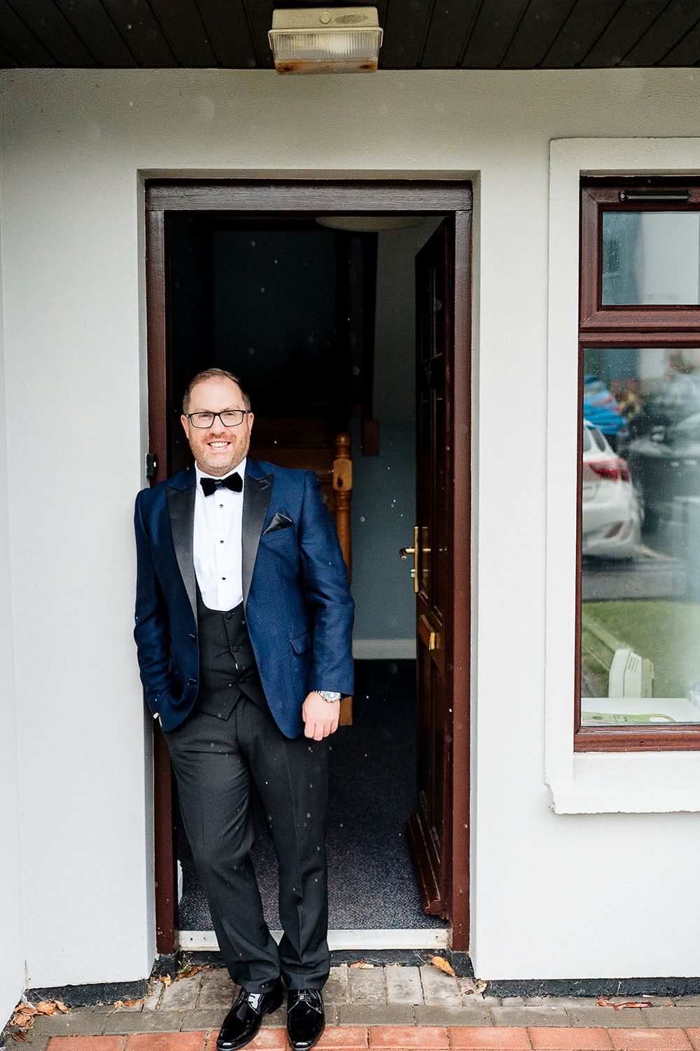 A person in a formal navy tuxedo and bow tie stands in a doorway of a white building.