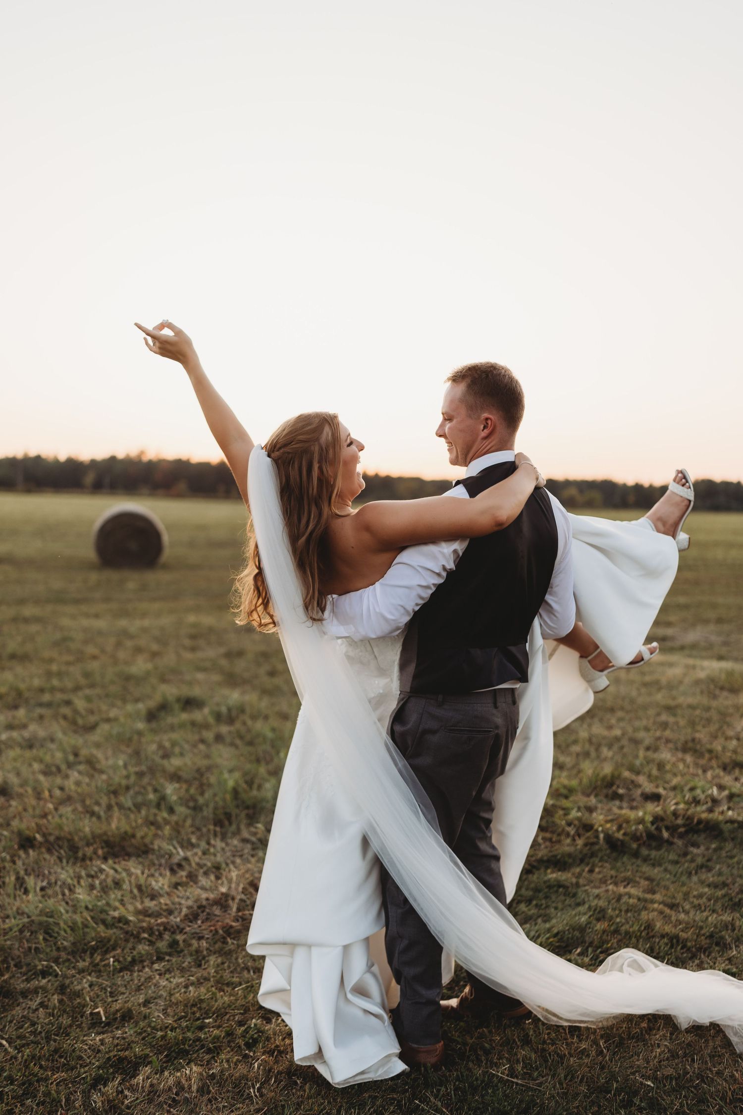 A couple dances in a field at sunset with hay bales in the background as the bride's white dress and veil flow in the breeze.