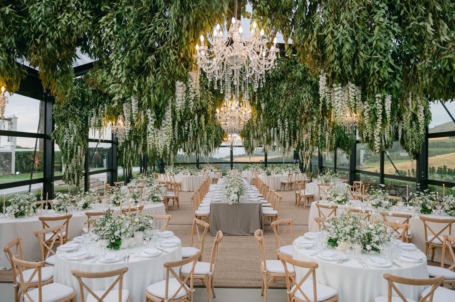 Elegant wedding reception setup with wooden cross-back chairs and white floral centerpieces under hanging greenery.