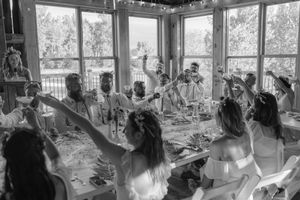 Wedding reception guests mingle and dance in a sunlit venue with large windows and rustic decor.