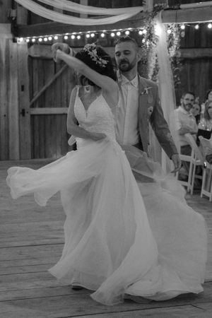 A couple in wedding attire dances together on a wooden deck under string lights in a romantic black and white photo series.