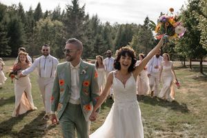 Group in white dresses and sage green attire pose joyfully outdoors among evergreen trees.