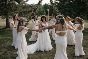 Wedding party dances in a circle on grassy field surrounded by pine trees.