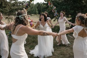 Wedding party forms a playful dance line outdoors in white dresses against a natural backdrop.