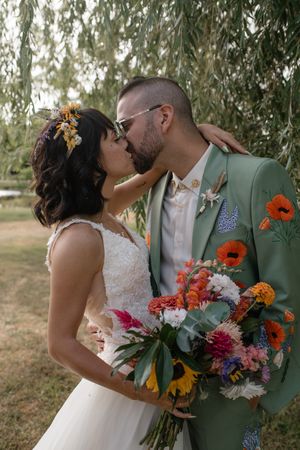 Romantic wedding couple embracing under willow trees with colorful floral bouquet.