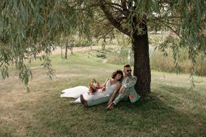 A couple relaxes together under a weeping willow tree on a grassy hill during a sunny summer day.