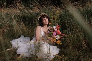 Bride in white dress sitting among tall grass with autumn flowers.