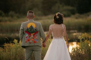 Newlywed couple holding hands at sunset with groom wearing custom painted suit jacket in outdoor setting.