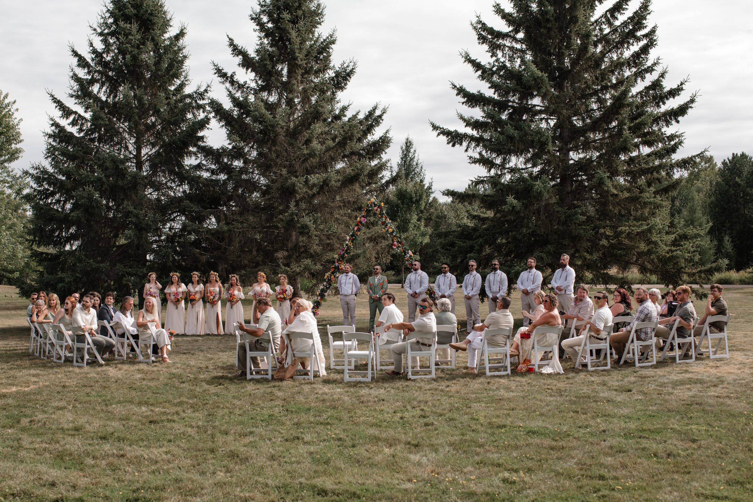 Large wedding party in white dresses and gray suits pose outdoors with evergreen trees in background.