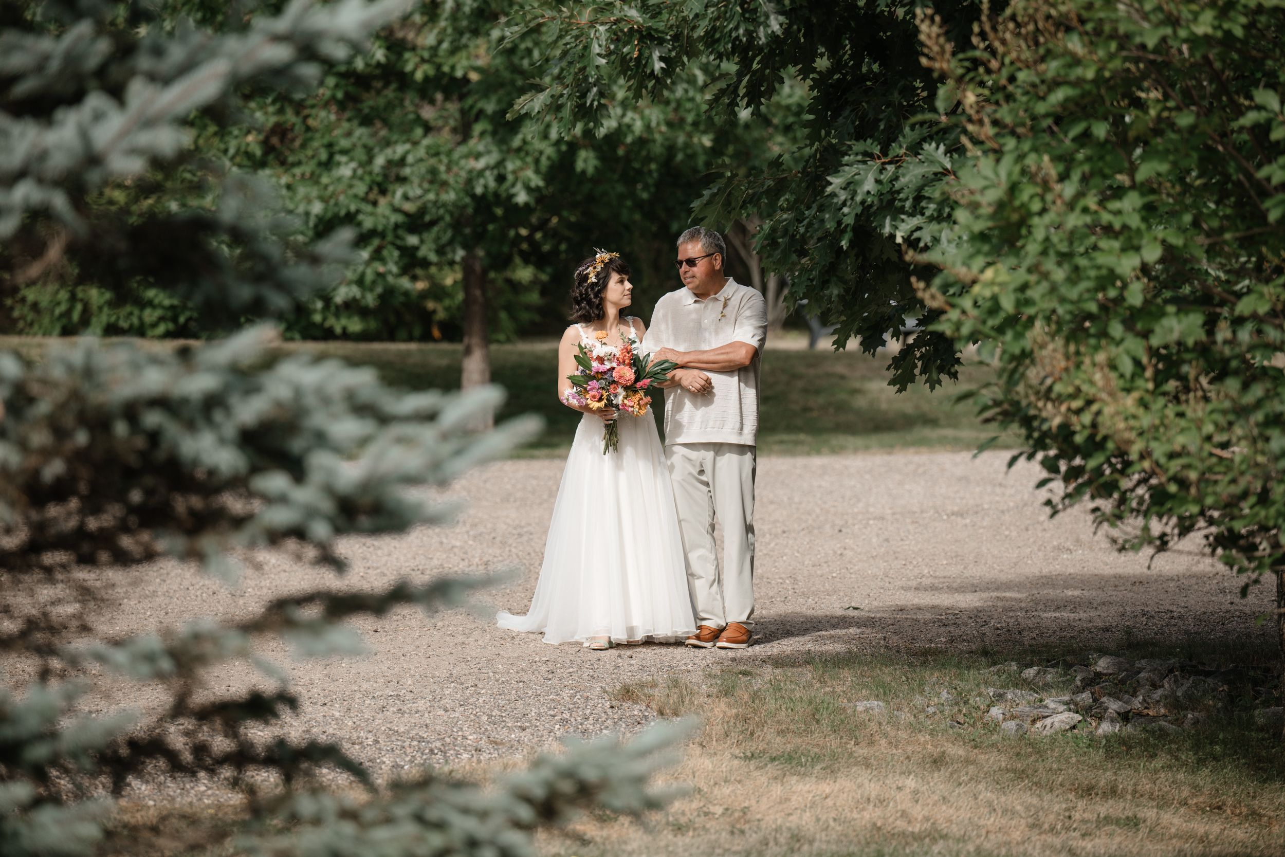 Wedding party walking along a tree-lined path in white dresses on a summer day.