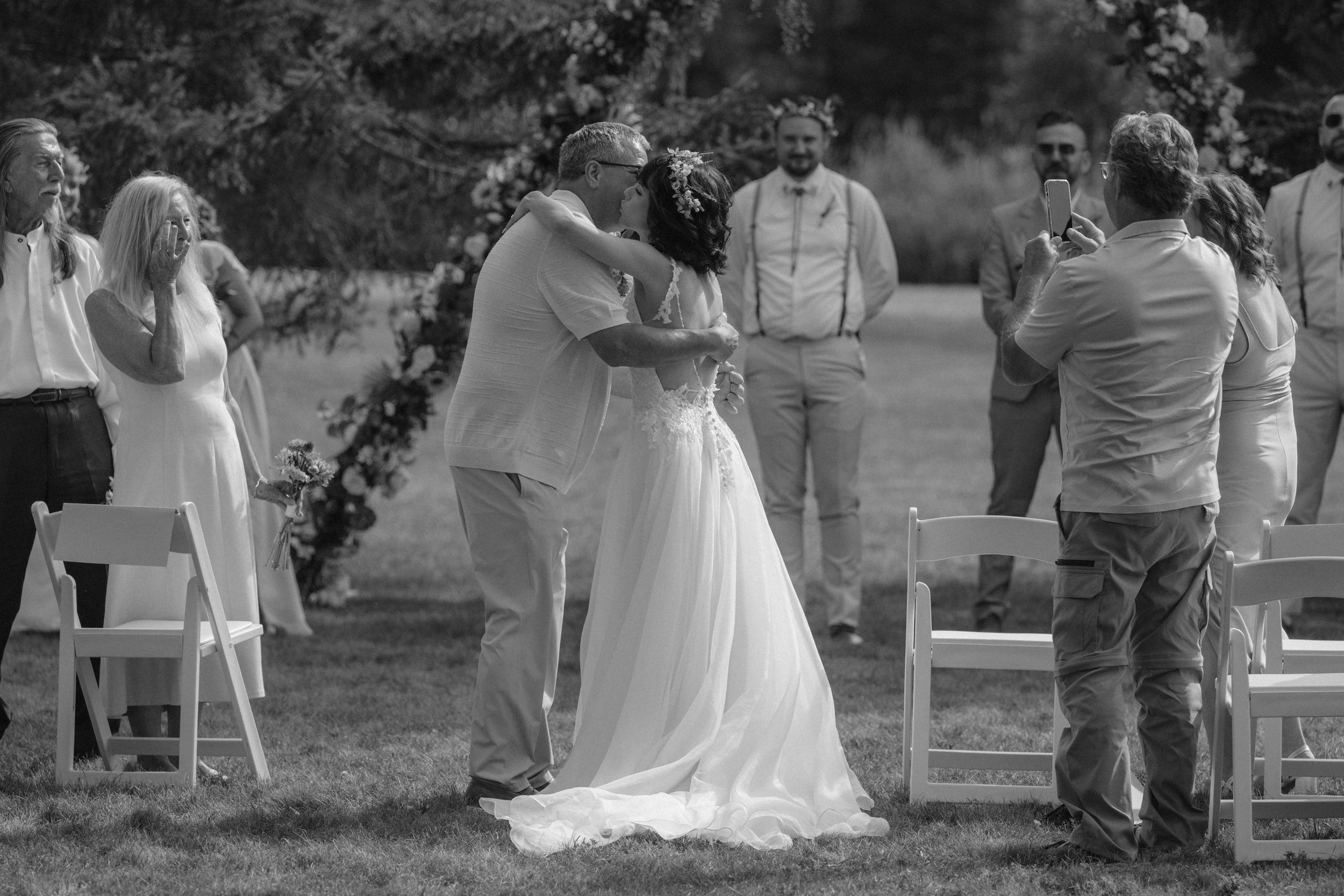 Wedding ceremony setup outdoors with white chairs and floral arrangements under tall evergreen trees.