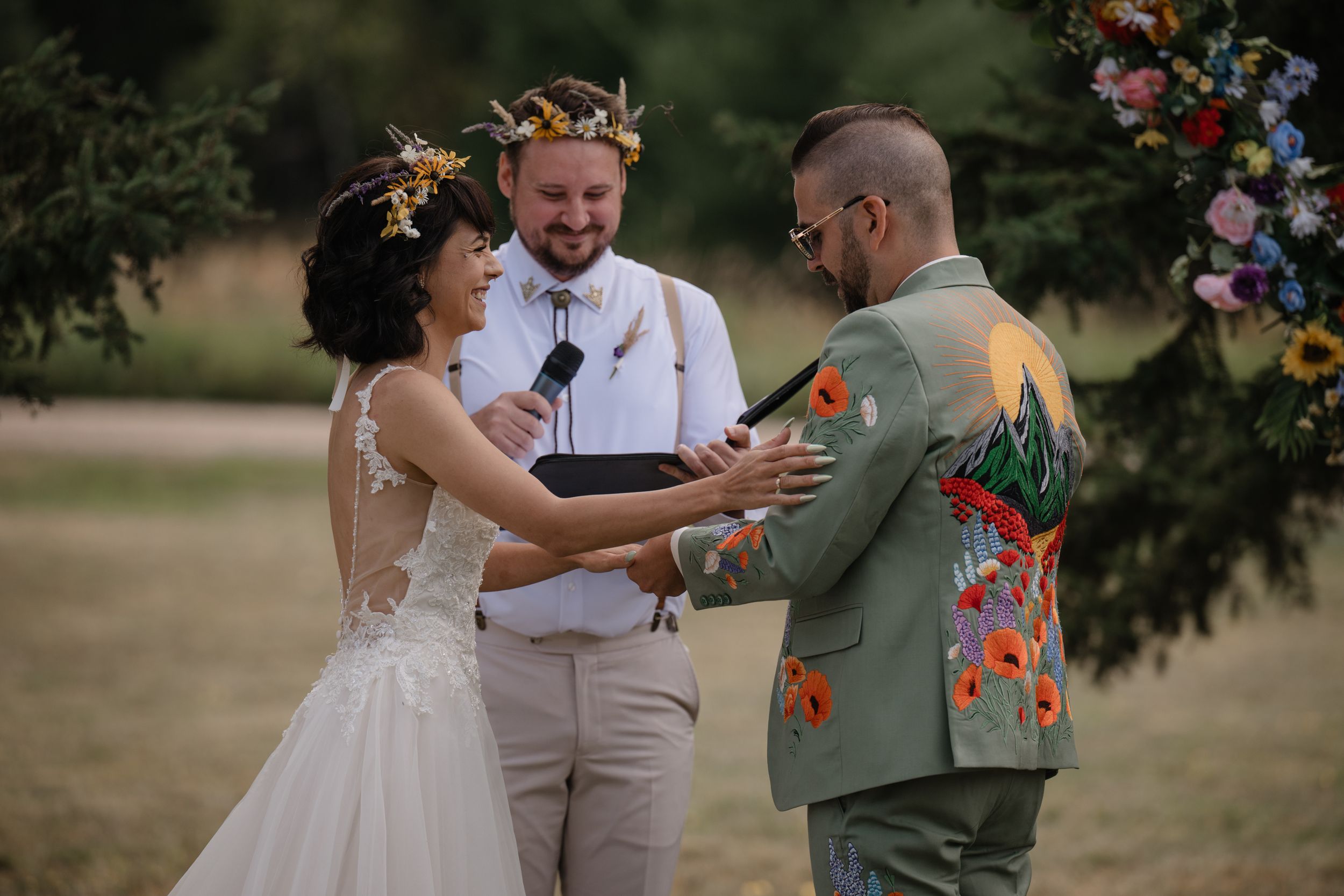 Wedding ceremony taking place outdoors with floral arch and groom in unique painted suit jacket.