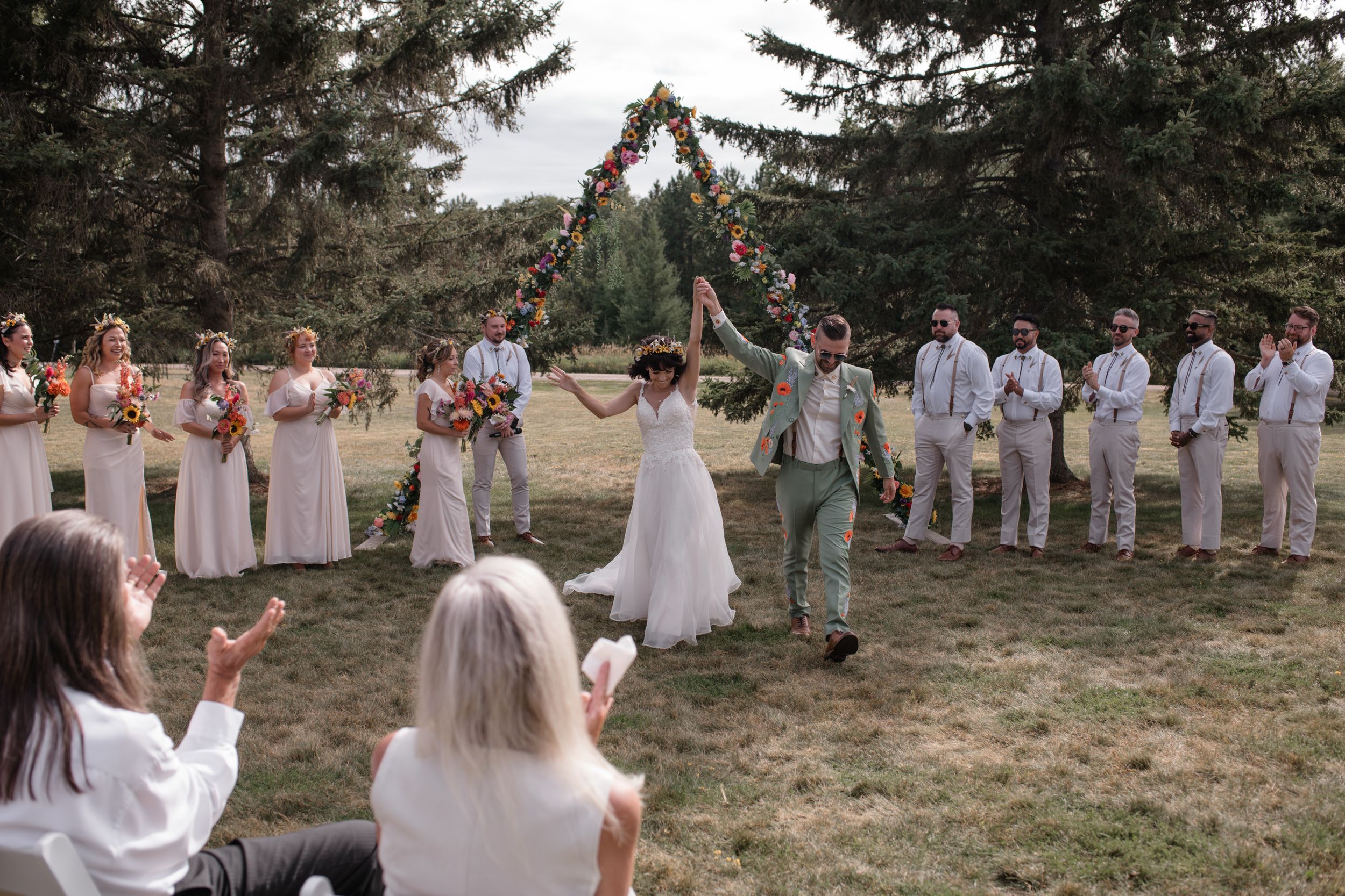 Wedding party celebrates outdoors in a line formation with coordinated dresses and attire against a natural backdrop.