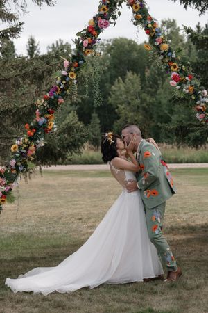 A romantic wedding couple shares a kiss under a floral arch decorated with vibrant flowers in an outdoor garden setting.