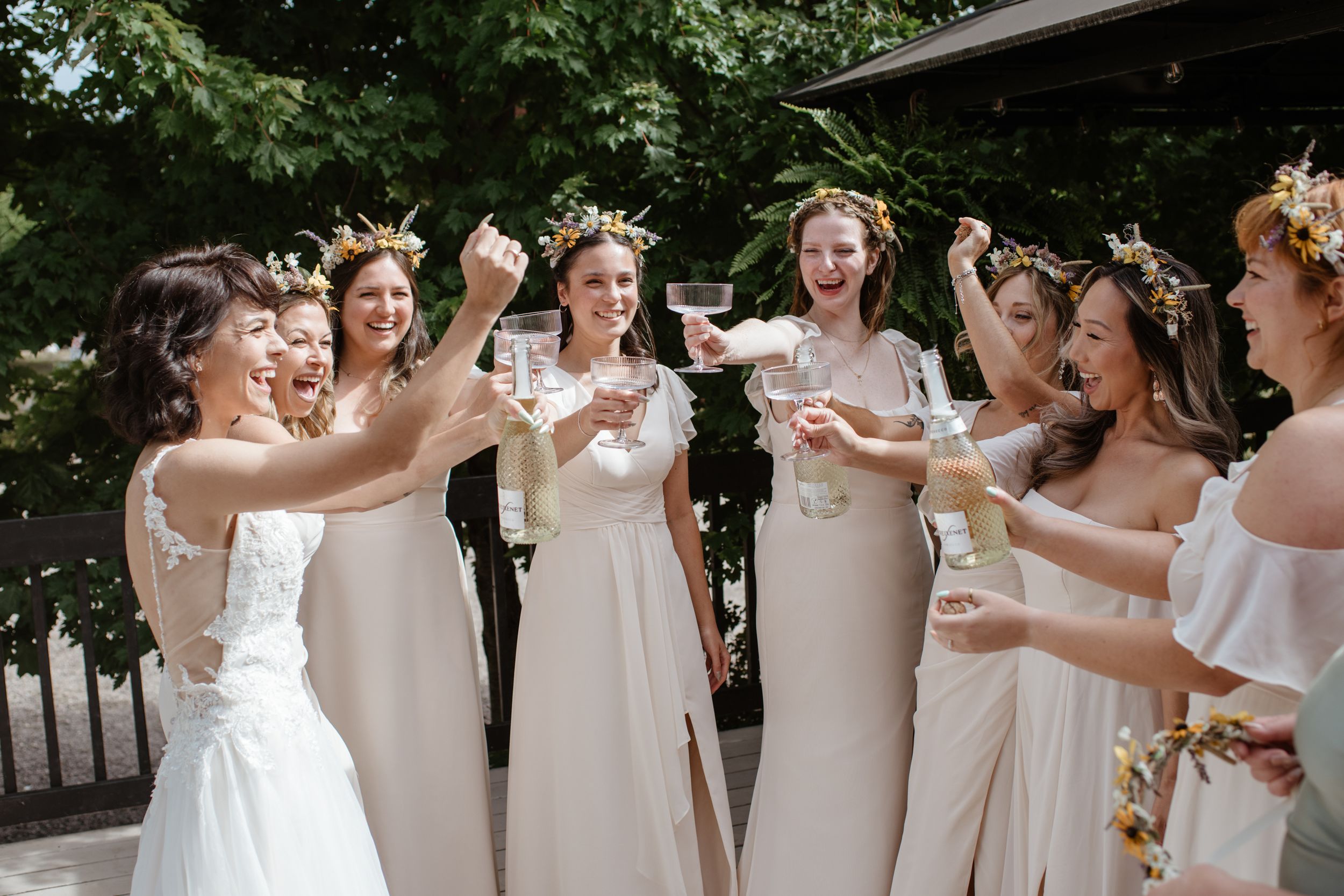 Bridesmaids in white dresses standing together outdoors under green trees on a sunny day.