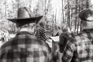 A black and white photograph shows people in plaid shirts and cowboy hats gathered outdoors in a wooded setting.