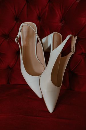 White leather slingback heels with pointed toes displayed against a deep red textured background.