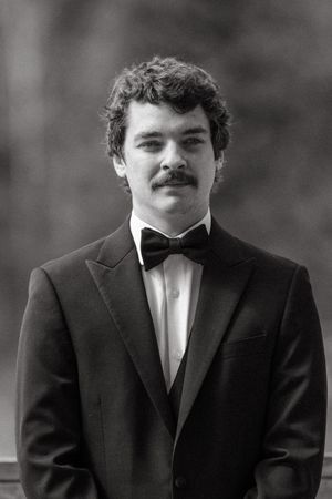 A formal portrait of a groom in a black tuxedo and bow tie with curly hair and a mustache.