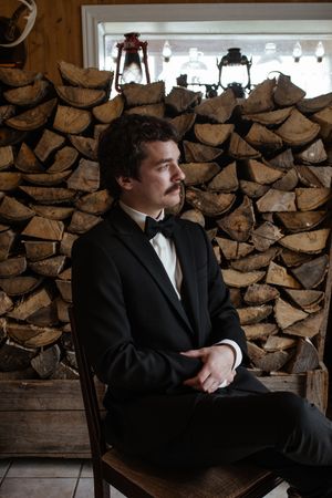 A figure in a dark suit sits thoughtfully against a backdrop of stacked firewood logs in a rustic setting.