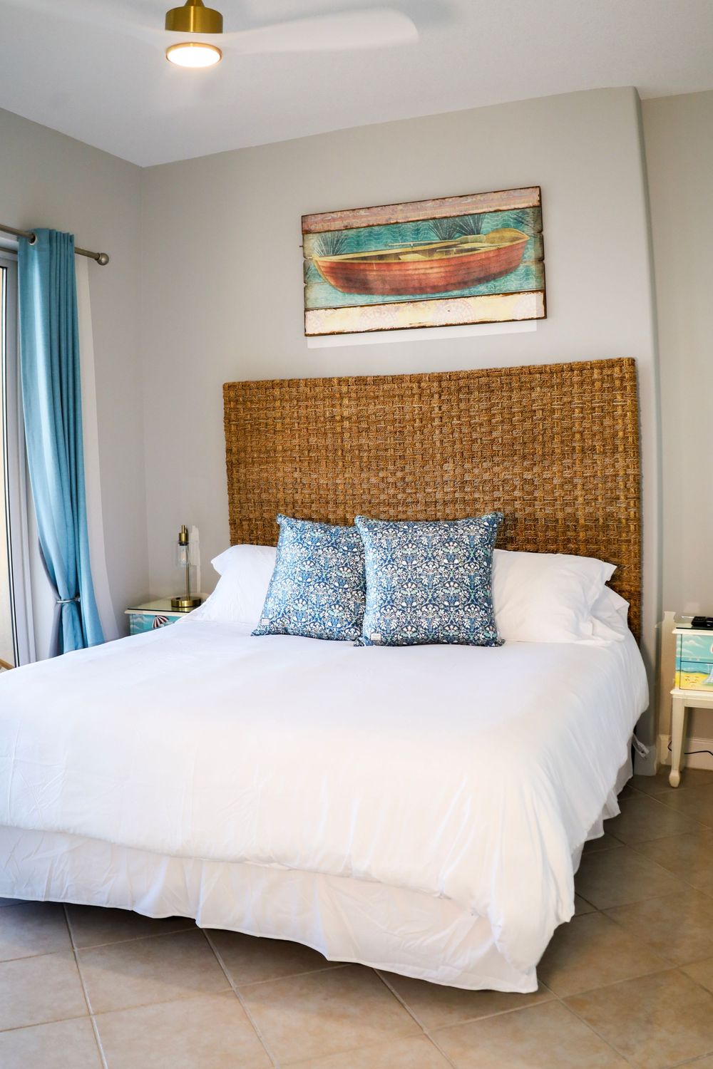 Coastal bedroom with woven headboard, turquoise curtains, white bedding, and wooden boat artwork above the bed.