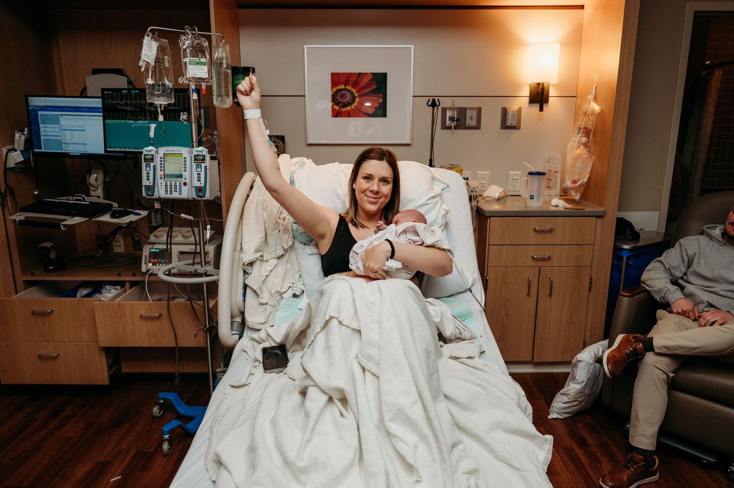 A mom raises her hand in a fist to celebrate her VBAC while holding her baby in a hospital bed.